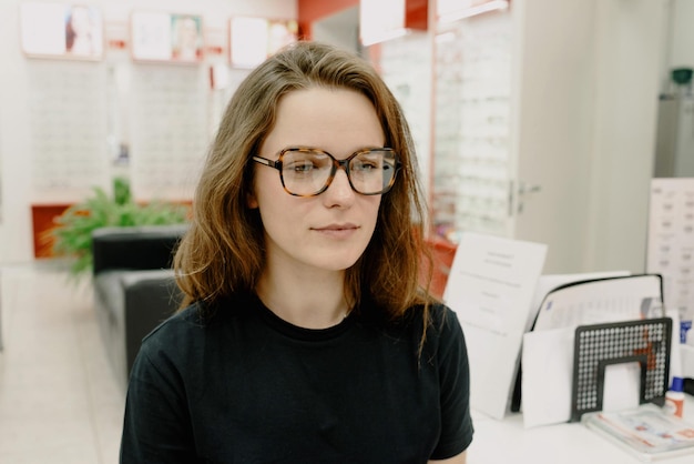 A woman wearing glasses sits in front of a desk with a display of eyewear.