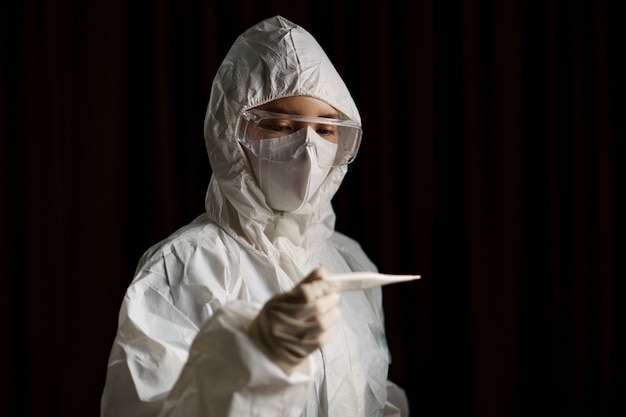 Woman wearing gloves with biohazard protective suit and mask holds thermometer