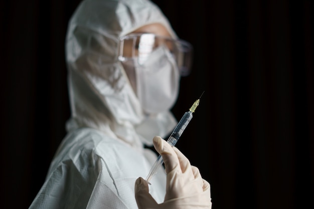 Женщина в перчатках с защитным костюмом и маской от биологической опасности держит вирусную вакцину со шприцем для профилактики и лечения от коронарного вируса (Covid-19)