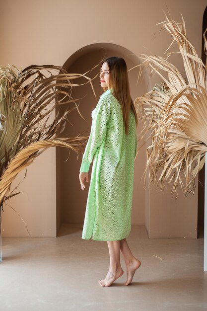 A woman wearing a green robe stands in front of a wall with a plant in the background.