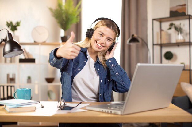 Woman wearing headphones sitting at the table at modern apartment and showing victory sign gesture