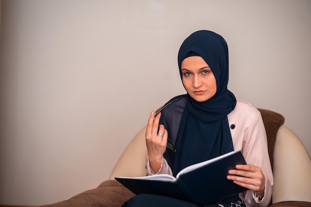 A woman wearing a hijab and glasses sits in a chair with a pen