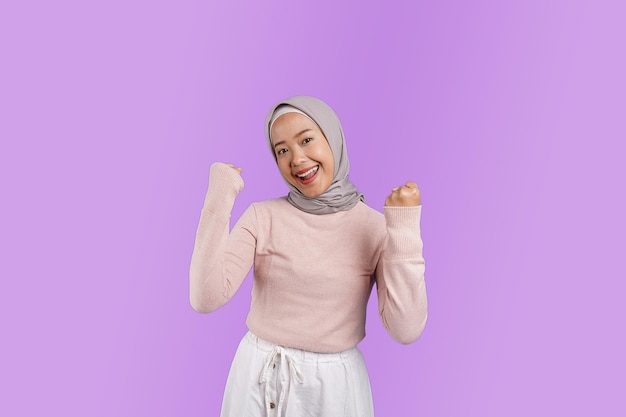 A woman wearing a hijab and a pink sweater smiles and raises her fist