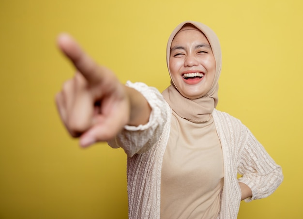 woman wearing hijab points to someone isolated on a yellow wall