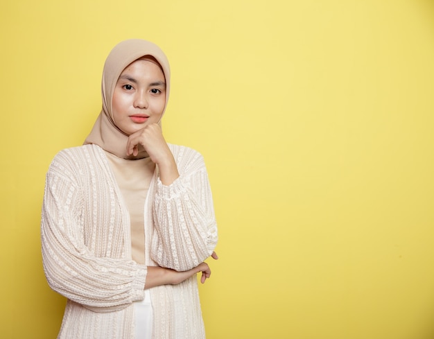 노란색 배경에 고립 된 카메라를보고 그녀의 턱을 잡고 웃는 hijab를 착용하는 여자