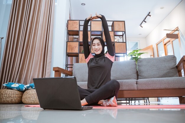 히잡 운동복을 입은 여성이 집에서 노트북을 보면서 손을 위로 뻗으면서 다리를 꼬고 앉아 있습니다.