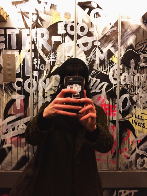 Foto donna con una giacca che si fa un selfie con lo smartphone contro un muro di graffiti