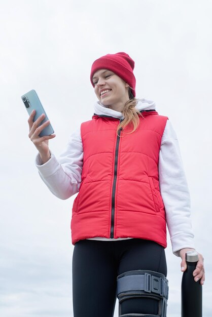 Woman wearing knee brace or orthosis after leg surgery walking\
in the park using smartphone