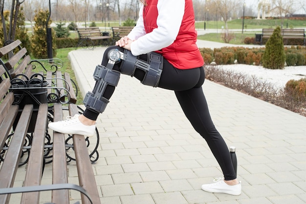 Woman wearing knee brace or orthosis after leg surgery walking\
in the park