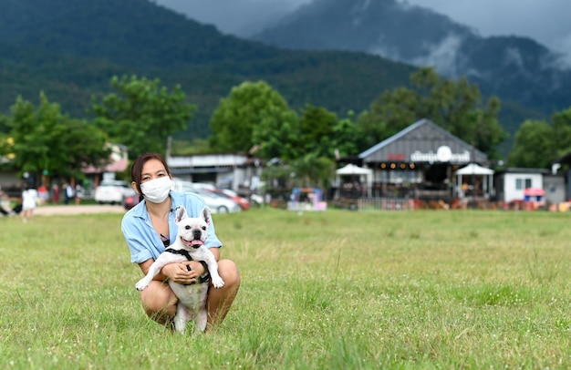 A woman wearing a mask hug a white French bulldog on the lawn looking at the camera, mountains and clouds behind.