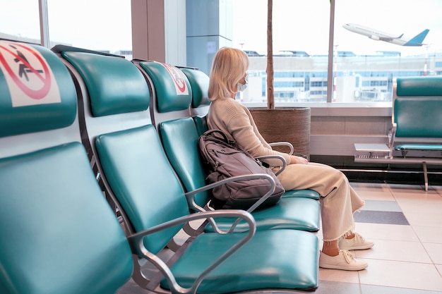 Woman wearing mask looking through window and sit social distancing chair at airport. Travel vacation business trip concept. Selective focus.