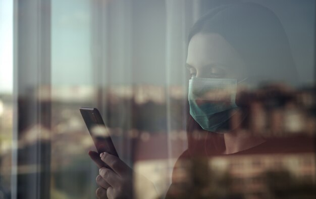 Woman wearing mask and using smartphone behind closed window during quarantine