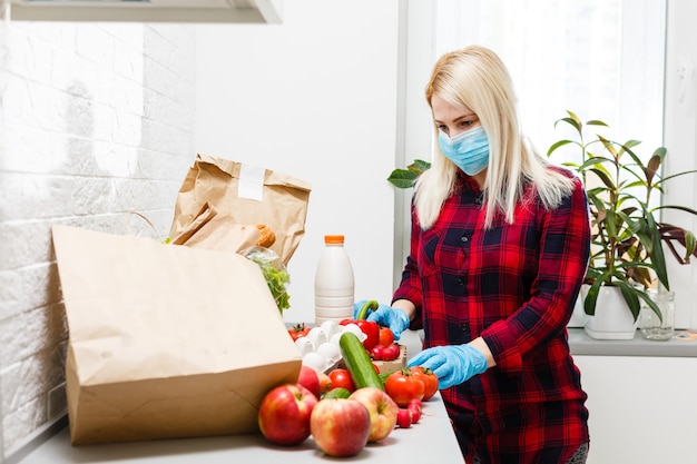 woman wearing medical face mask Disassembles food bags at home in the kitchen. quarantine. health concept. Corona Virus. order of products online. Delivering products to home