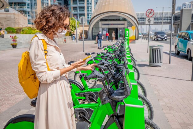 A woman wearing a medical mask uses a smartphone app to rent a shared electric bike to travel around the city to avoid dangerous contact with people during the coronavirus pandemic