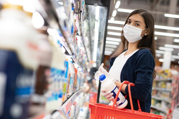 Woman wearing medical mask with shopping basket choosing daily milk from shelf at grocery