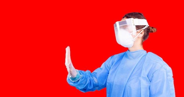 Woman wearing medical uniform in gloves and protective plastic mask showing with her hand stop coron...
