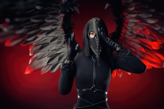 Photo the woman wearing a plague doctor mask with leather straps and black wings on a dark red background