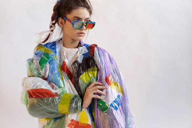 Woman wearing plastic on white wall. female model in clothes\
and shoes made of garbage. fashion, style, recycling, eco and\
environmental concept. too much pollution, we\'re eating and taking\
it.