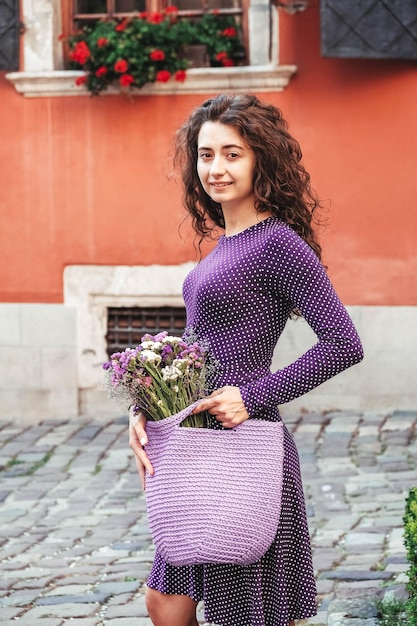 Woman wearing purple polka dot dress holding knitted bag with flowers posing in street of city