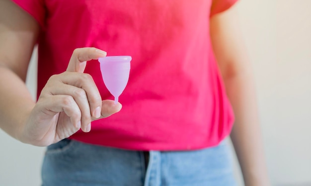 白い背景の前でピンクの月経カップを握っている赤いシャツを着た女性