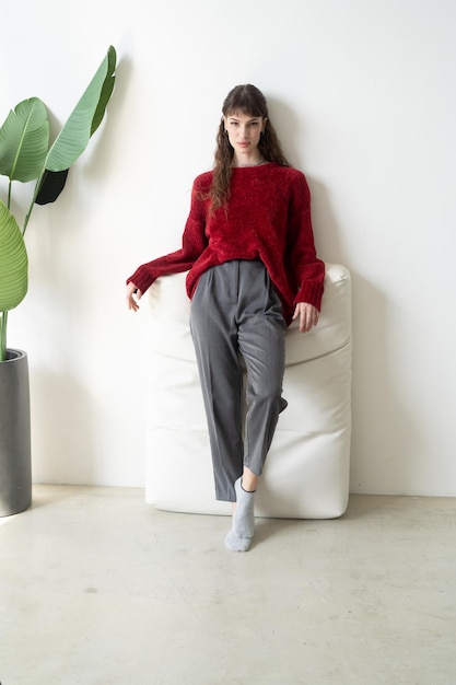 A woman wearing a red sweater and grey pants sits on a white couch.