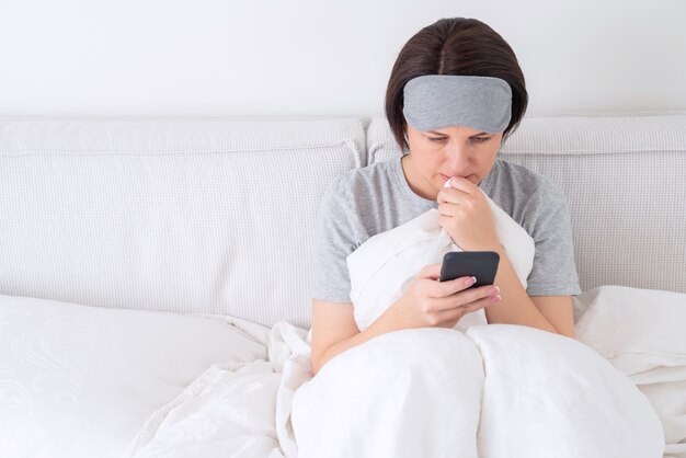 彼女は布団で覆われたベッドに座っているようにスマートフォンで寝ているマスクテキストメッセージを着ている女性