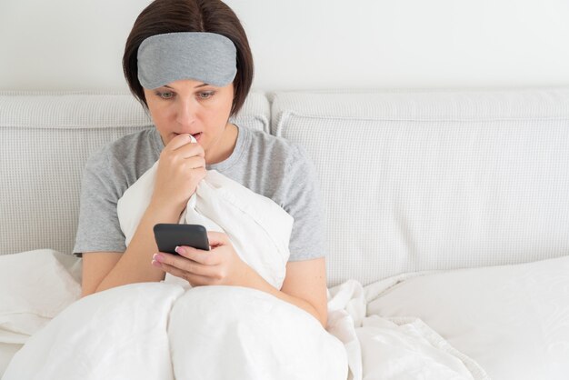 Woman wearing sleeping mask texting on smartphone as she sits in bed covered with duvet