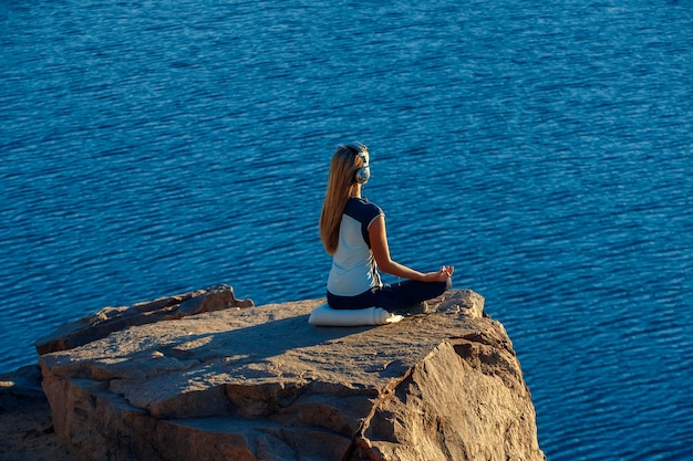 Donna che indossa tuta sportiva e cuffie seduto nella posizione del loto su roccia sopra il mare, meditando, ascolta musica. yoga all'aperto.