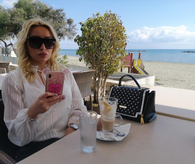 Фото Женщина в солнцезащитных очках использует смартфон, сидя в ресторане на открытом воздухе