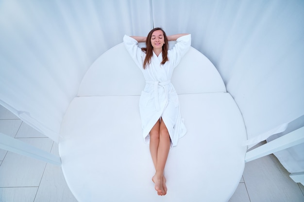 Woman wearing white bathrobe with closed eyes and hands behind head lying on a lounger and enjoying relaxing time and feeling good at a wellness spa resort. Easy lifestyle and satisfaction