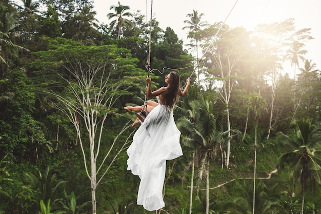 Woman wearing white dress swinging on rope swings with beautiful view on rice terraces and palm trees in the Bali Island