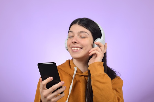 A woman wearing a yellow hoodie is holding a phone and wearing headphones and listening to music