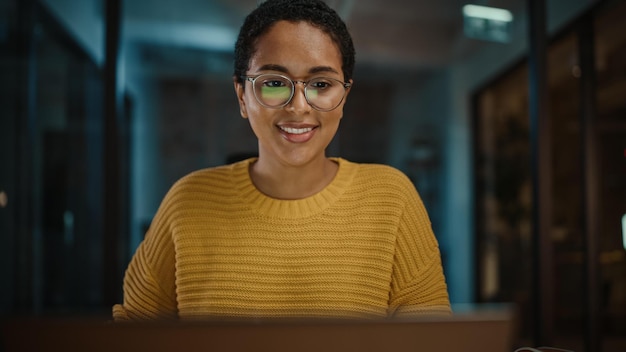 A woman wearing a yellow sweater smiles at the camera while looking at a laptop.