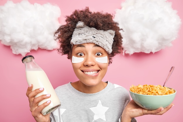 woman wears pajama sleepmask patches under eyes holds bowl of cereals and milk has nutrient breakfast poses indoor