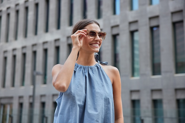  woman wears stylish sunglasses blue dress looks away happily poses on blurred city building enjoys spare time