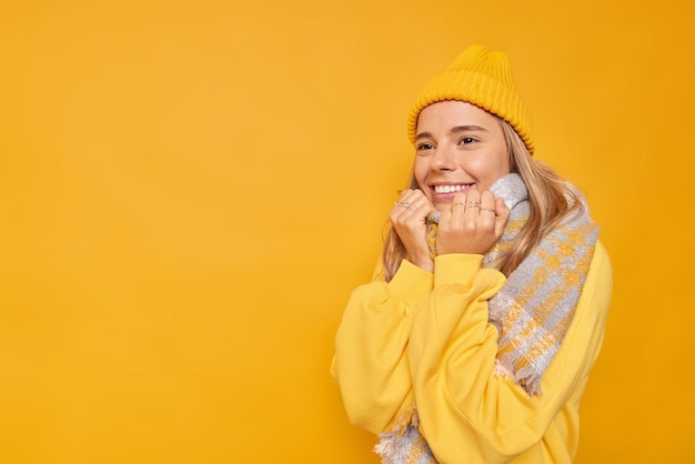 woman wears warm scarf around neck smiles gladfully feels good wears hat and casual jumper poses against vivid yellow blank empty space for your information.
