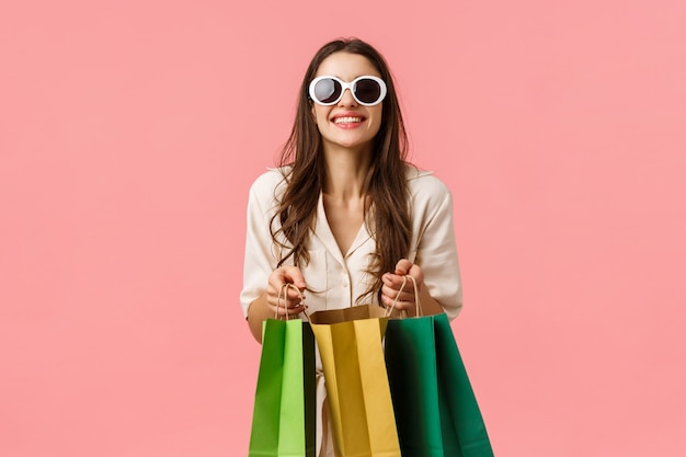 Woman went shopping, buying new clothes, special discounts for holiday season, standing with shop bags and smiling, wearing sunglasses, standing pink wall excited