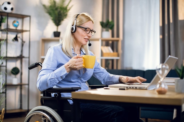 Woman in wheelchair holding cup of coffee and using laptop