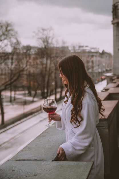 Woman in a white bathrobe with a glass of wine looking from the balcony
