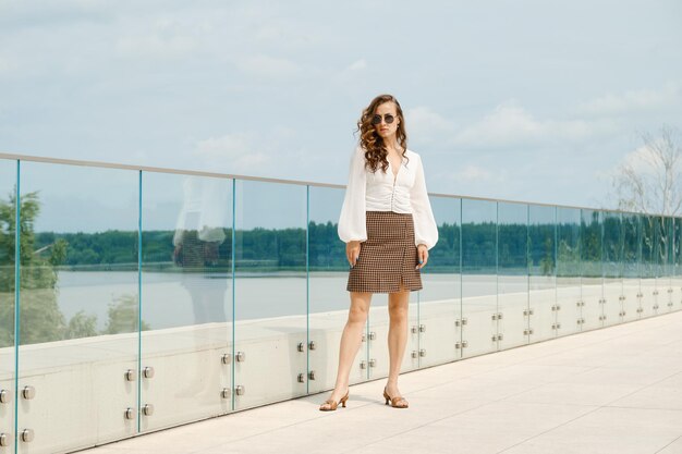 Woman in white blouse and brown mini skirt standing near glass railing of balcony