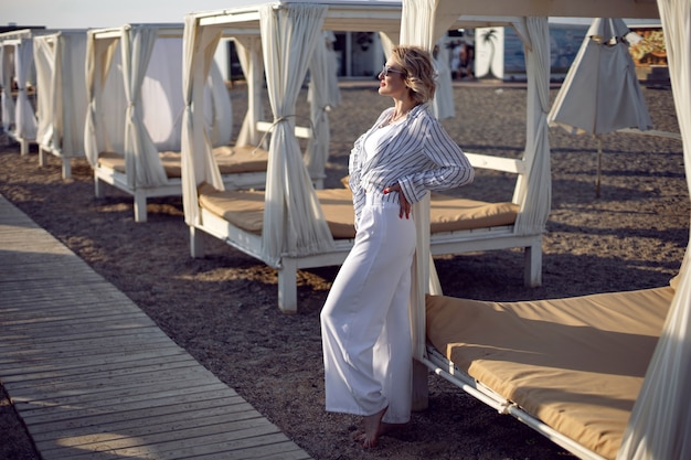 La donna in abiti bianchi e occhiali da sole è in piedi sulla spiaggia vicino al legno