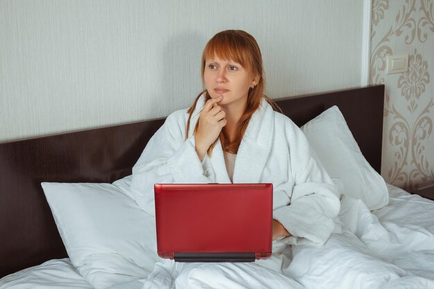 A woman in a white coat in bed with a laptop is thinking and looking into the distance