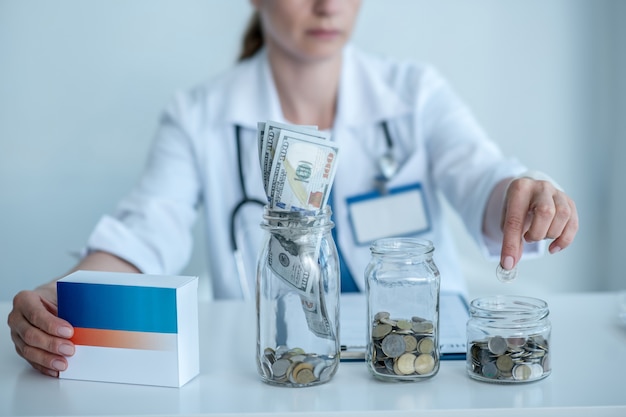 Woman in white coat sitting at the table with jars with coins