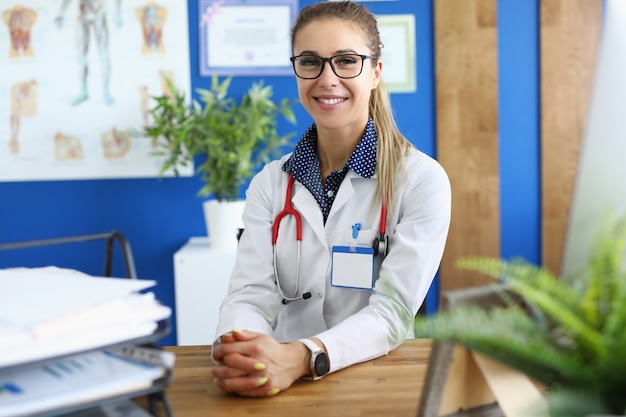 Woman in white coat and with red stethoscope around her neck sit with her hands folded at table.