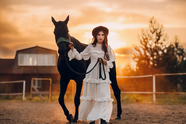 Woman in white dress and black hat with her horse at sunset outdoors ranch