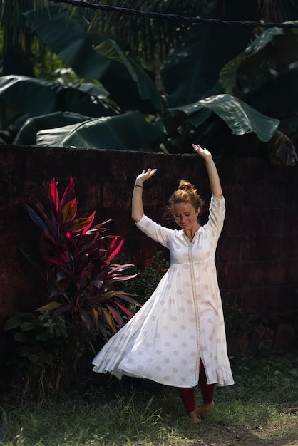 A woman in a white dress dances in front of a tropical plant