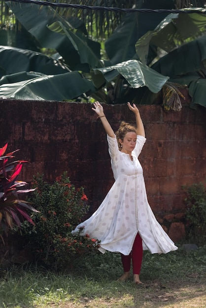 A woman in a white dress dances in front of a wall with tropical plants.