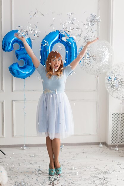 A woman in a white dress is standing in front of balloons with the number 30 on it.