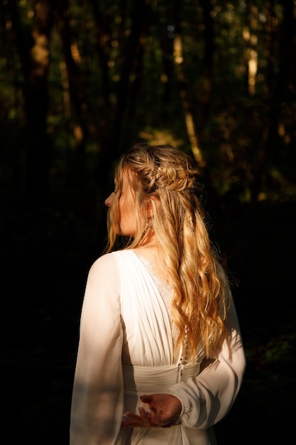 A woman in a white dress stands in the woods, looking back at the sun