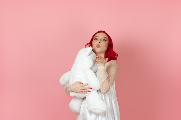 woman in a white dress and with red hair blows a kiss and hugs large white teddy bear